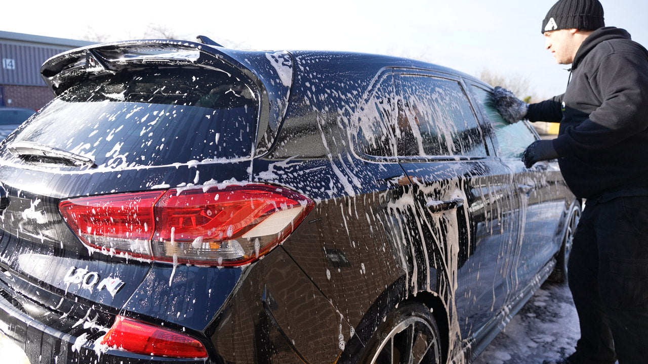 Black car covered in shampoo foam during a professional car wash, demonstrating safe washing techniques to help prevent paint scratches.